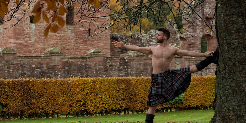 The Kilted Yogi demonstrates a pose against a tree, in front of a castle